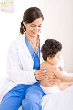 A female doctor in a white coat uses a stethoscope to listen to a young childs back. The child, wearing only a diaper, sits on an exam table facing away from the doctor.