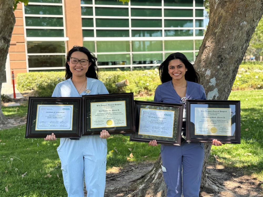 Two people stand outside on grass, each holding two framed certificates. They are smiling, with trees and a building with large windows in the background. Both are wearing medical scrubs.