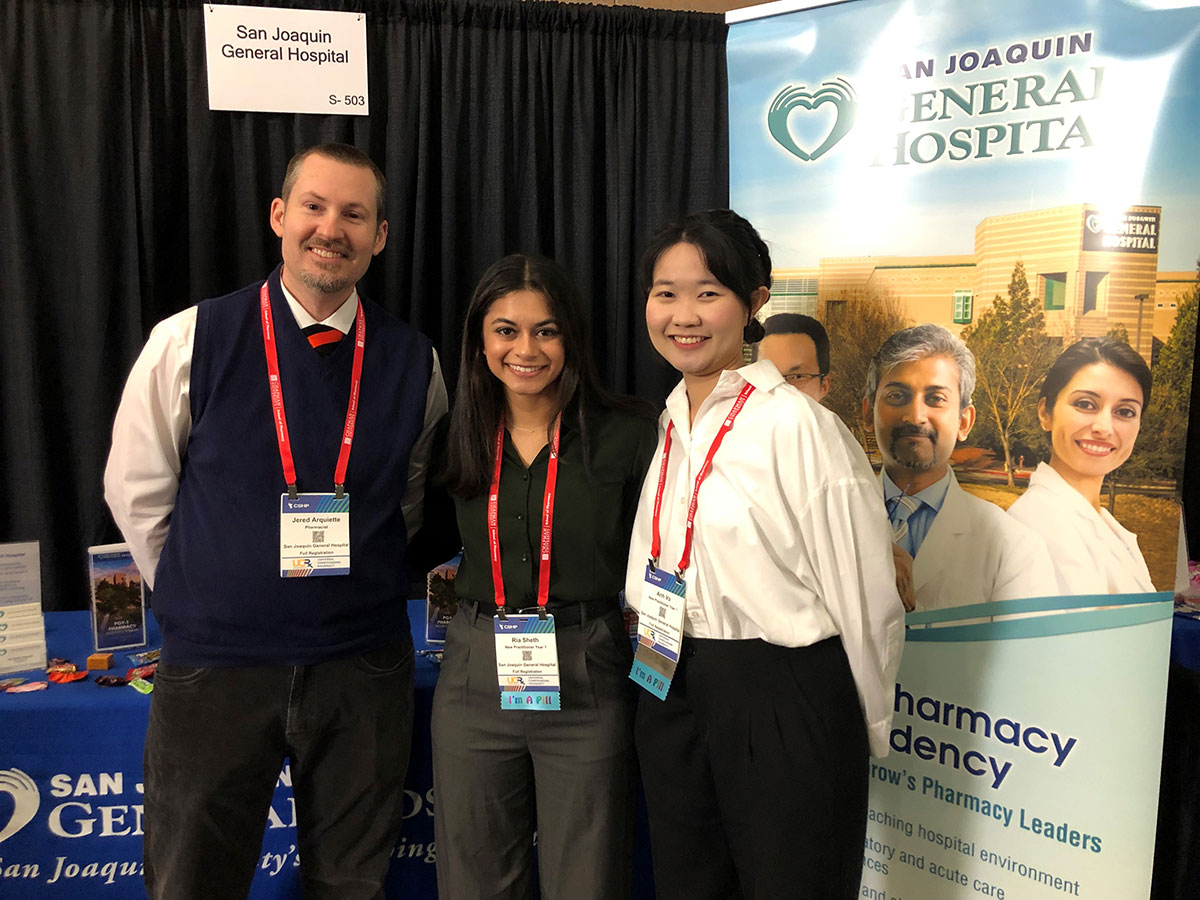 Three people stand smiling in front of a San Joaquin General Hospital booth at an event. They wear name badges and business attire, with a banner in the background promoting a pharmacy residency program.