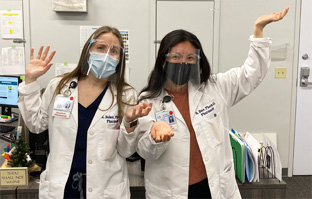 Two women wearing white lab coats, face masks, and face shields stand indoors with their arms raised and palms up, appearing to gesture or shrug in a medical or office setting.