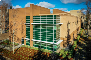 Two modern San Joaquin General Hospital buildings: the top one has a glass facade with beige vertical stripes and a covered entrance; the bottom one features large glass windows, metal beams, and trees in front, both photographed in daylight.