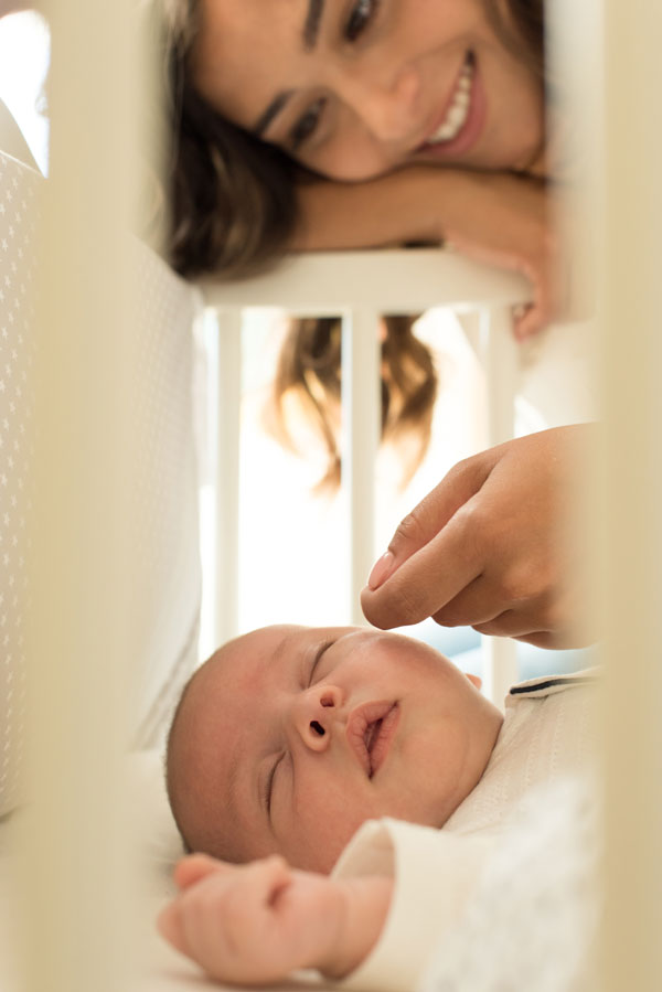 A smiling woman looks lovingly at a sleeping baby lying in a crib, gently reaching through the crib bars to touch the babys face.