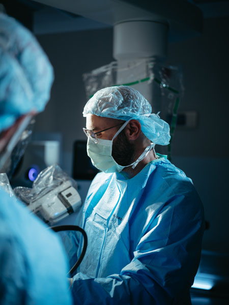 A surgeon wearing a surgical mask, hairnet, and gown is focused on performing a procedure in an operating room, with medical equipment and another medical professional visible nearby.
