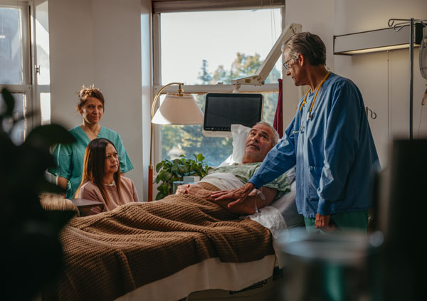 An older man lies in a hospital bed, covered with a brown blanket, while a doctor stands beside him touching his arm. Two women, possibly family and a nurse, stand nearby, all engaged in conversation in a bright hospital room.