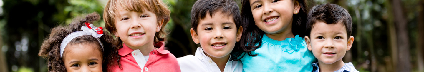 Five young children stand close together outdoors, smiling at the camera. They are dressed in colorful clothing, and a blurred background of trees suggests they are in a park or garden.